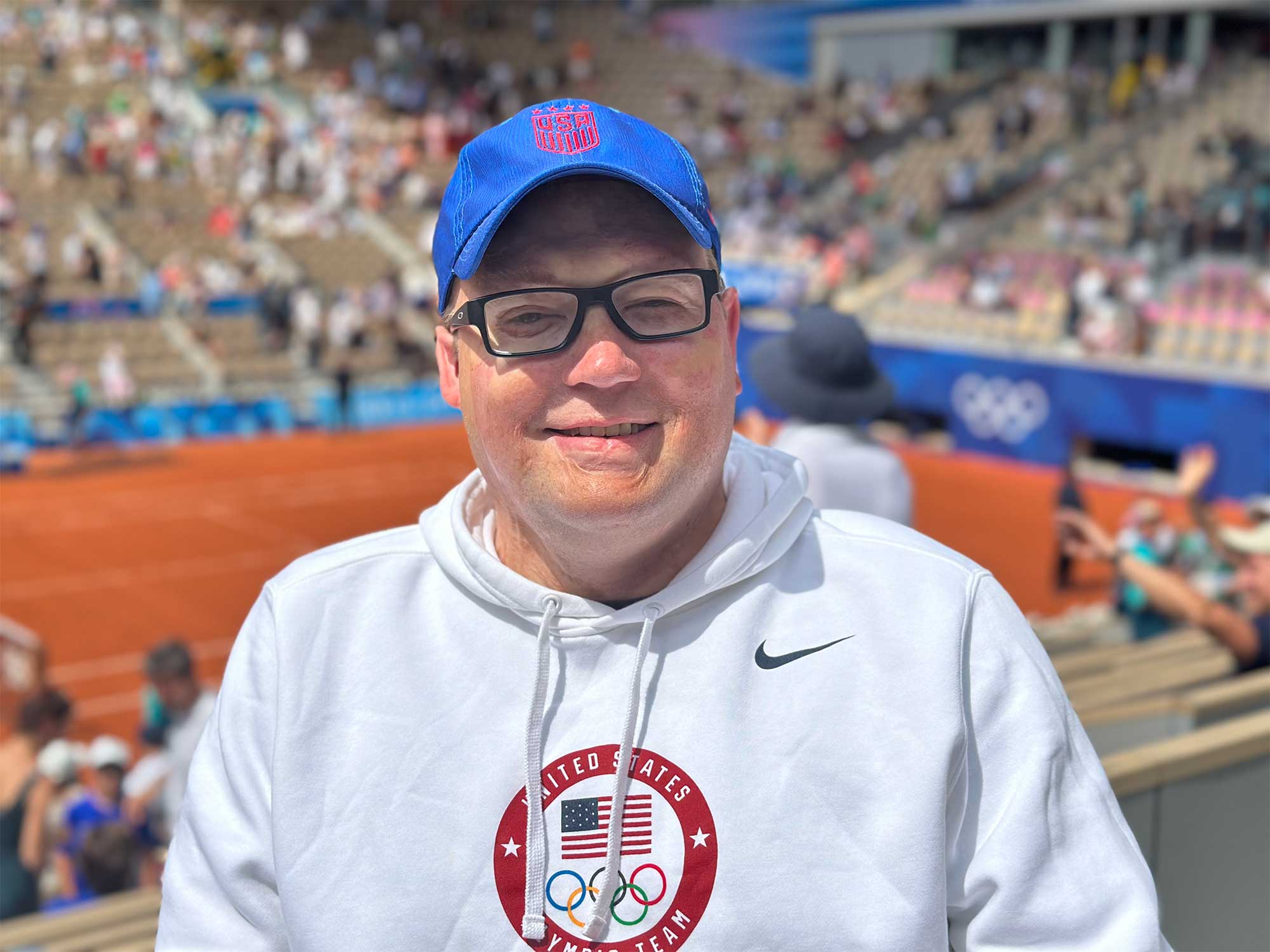 John wearing a white United States Olympic team hoodie with a blue USA baseball cap at a tennis stadium.