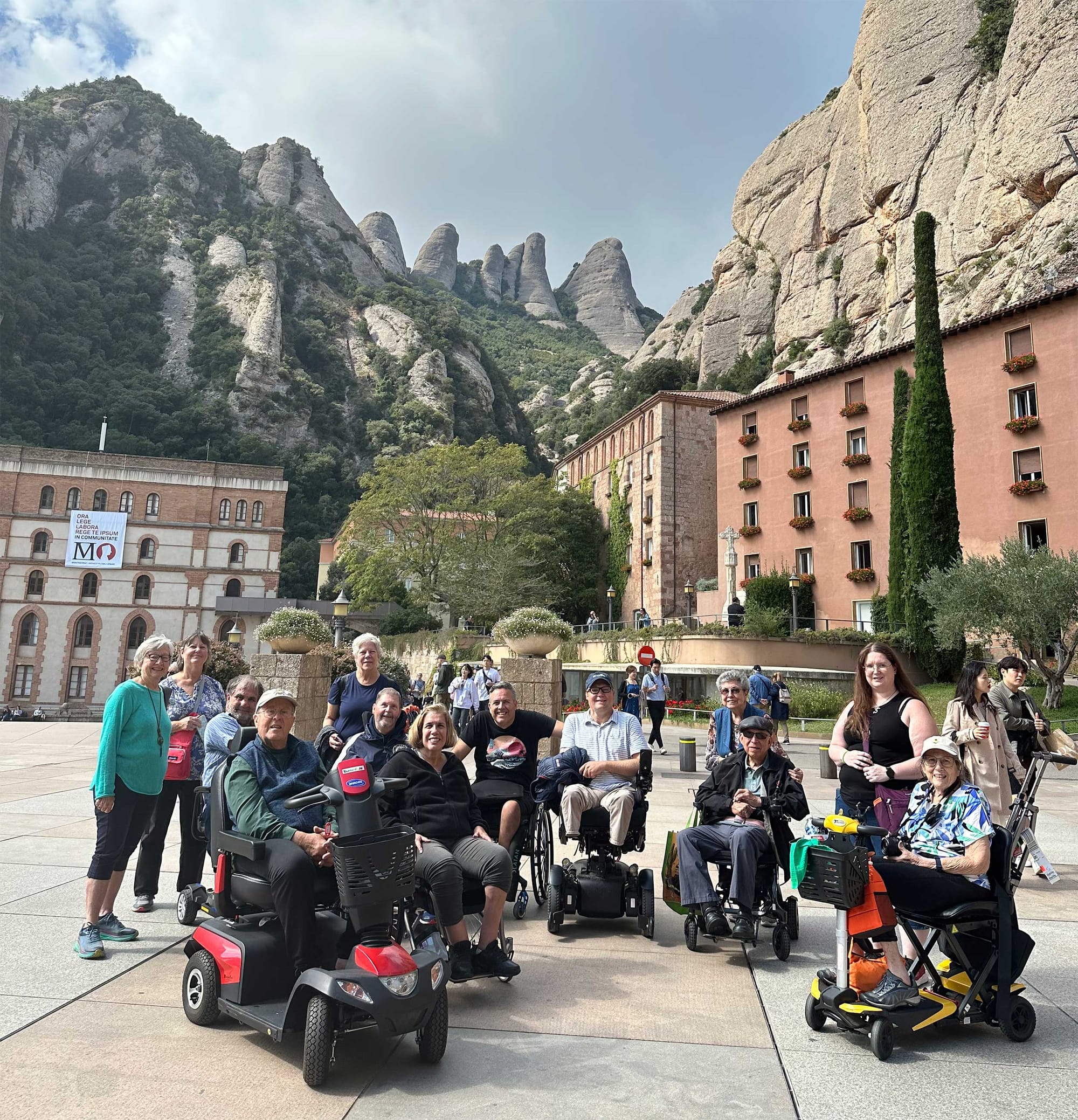 Group of wheelchair and scooter users at foot of large rock formation on mountainside.