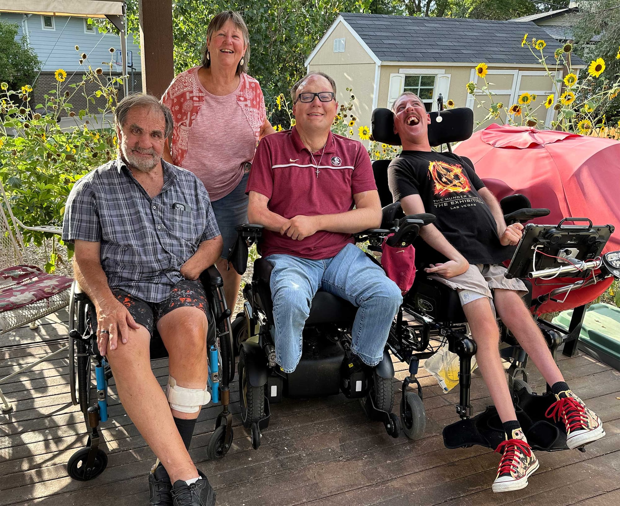 John pictured on a back porch with three wheelchair travel readers, surrounded by colorful sunflowers.