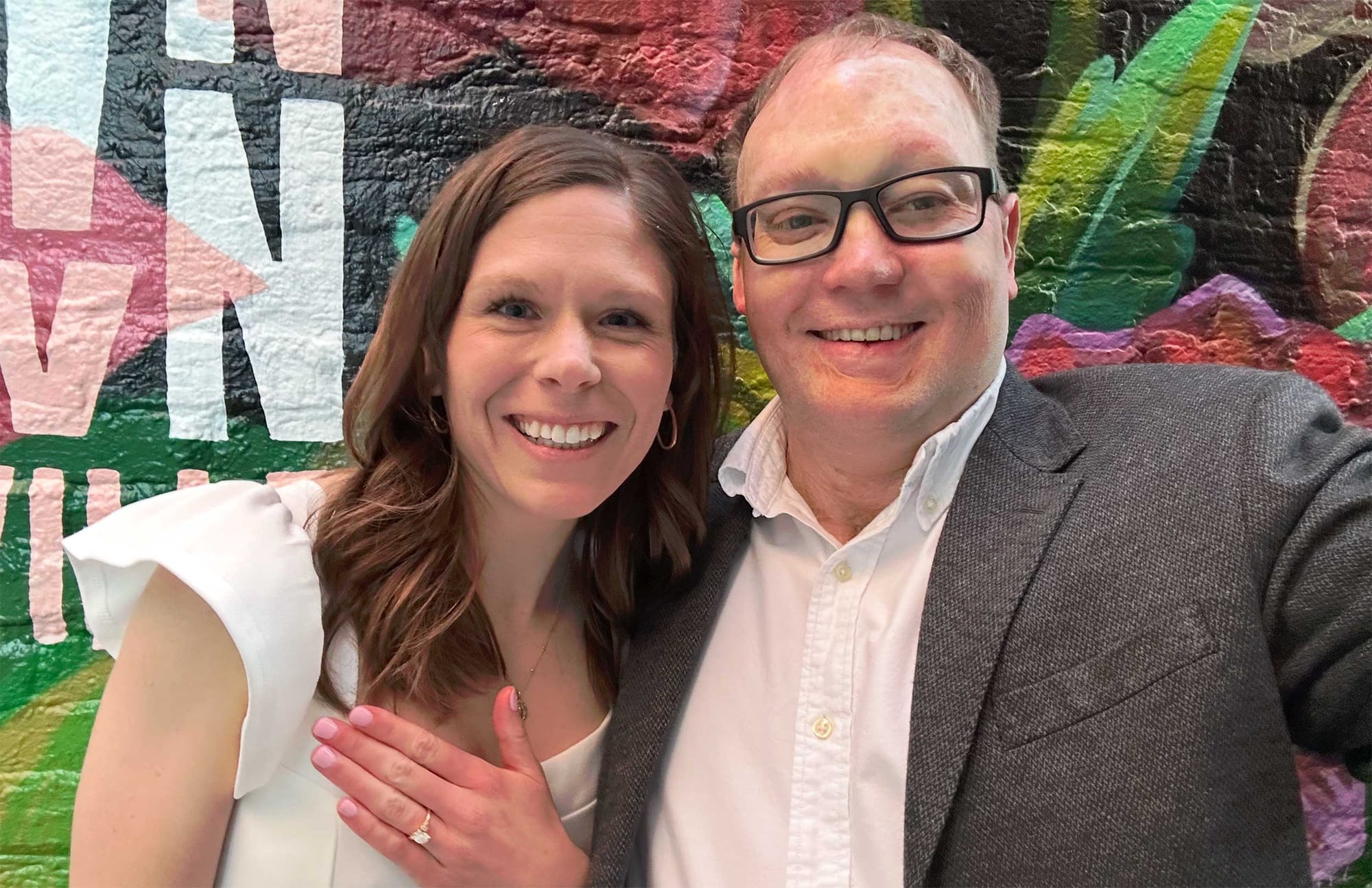 John taking a selfie with his fiancee, who is holding up her hand with an engagement ring.