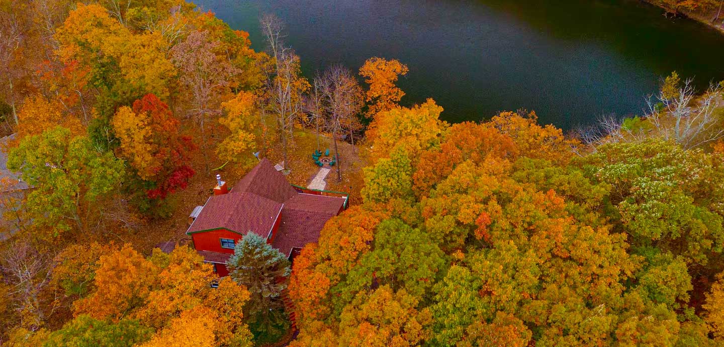 Aerial view of house in the middle of an autumn colored forest on a lakefront.