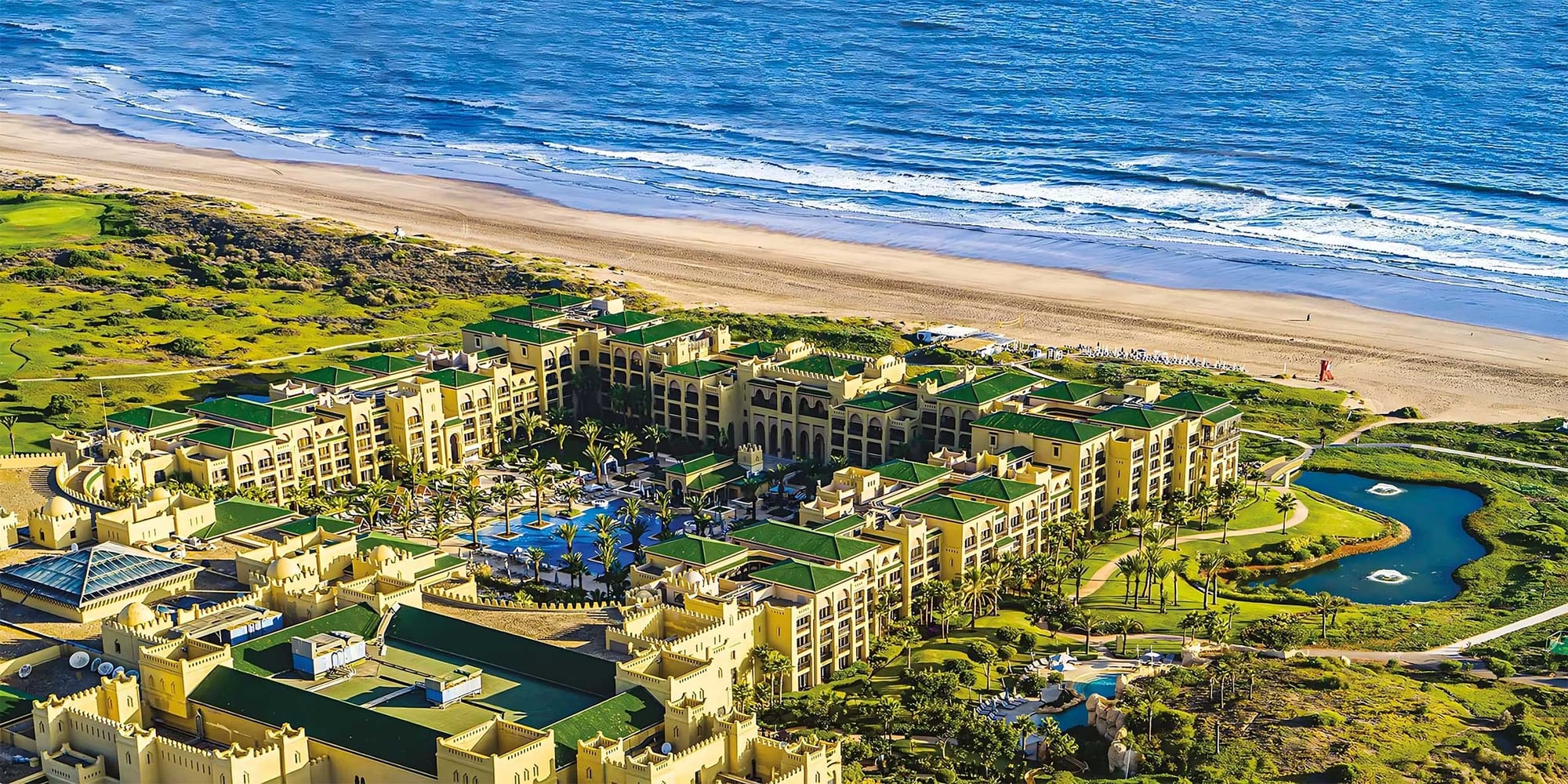 Aerial view of large beachfront resort with multiple towers, pools and a golf course.