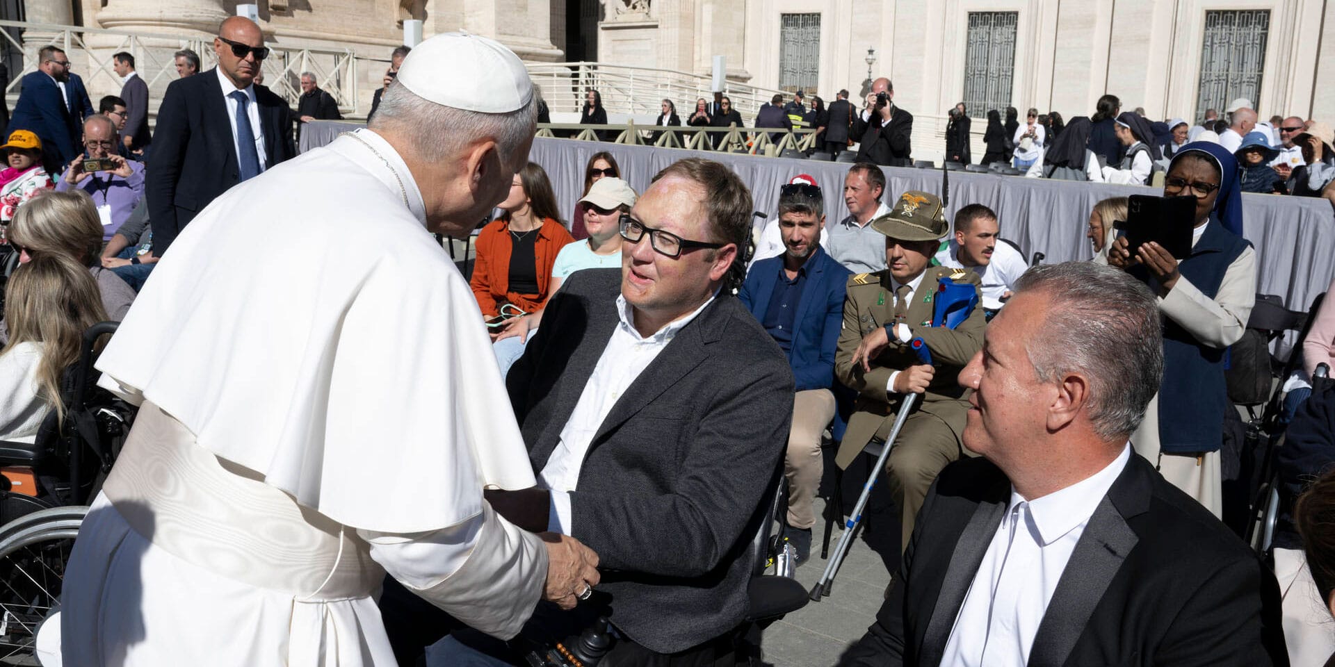 John seated in his power wheelchair shaking hands with Pope Leo.
