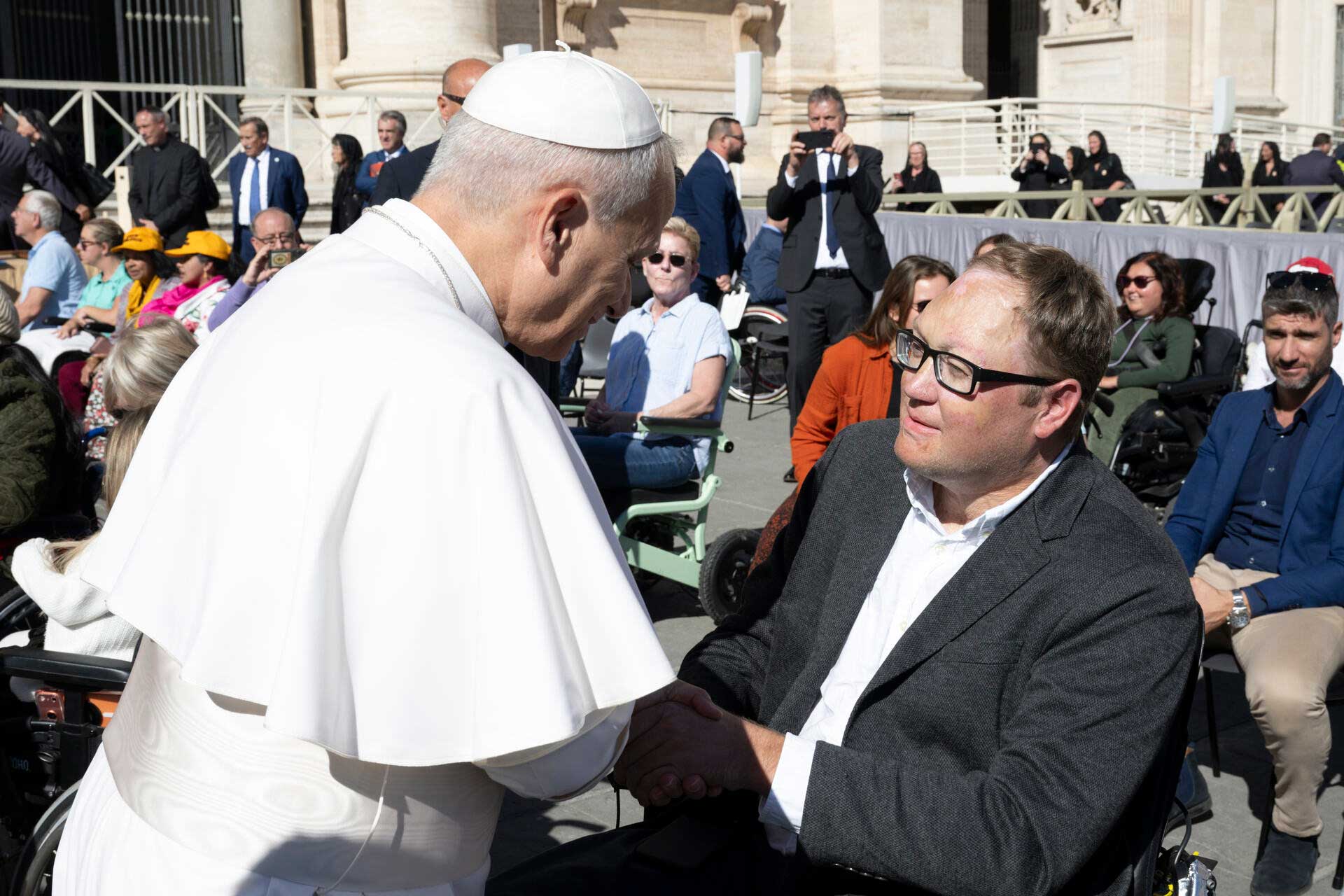 John seated in his wheelchair shaking hands with Pope Leo XIV in Vatican City.
