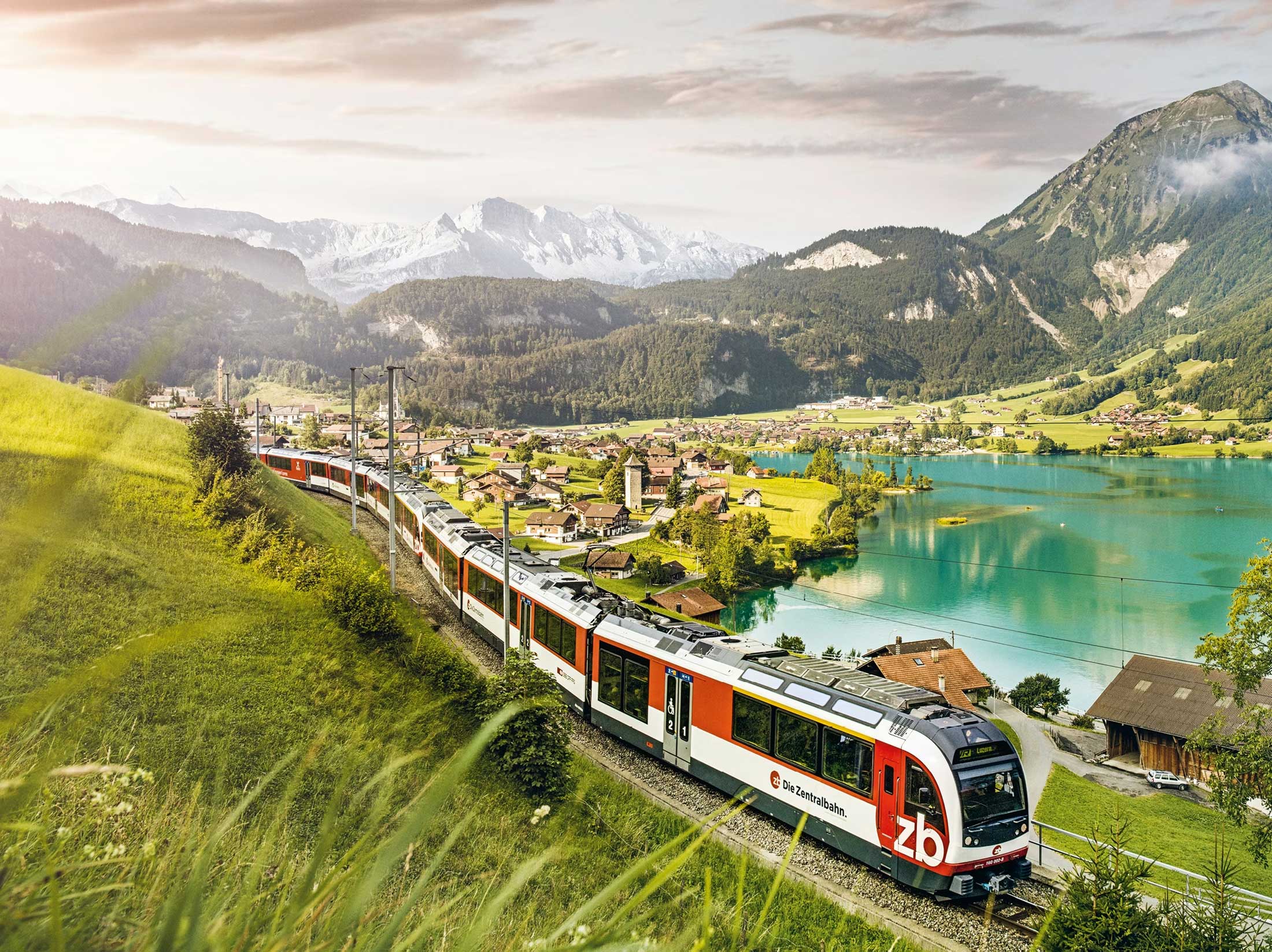 Train passing through scenic valley alongside lake and with mountains in the distance.