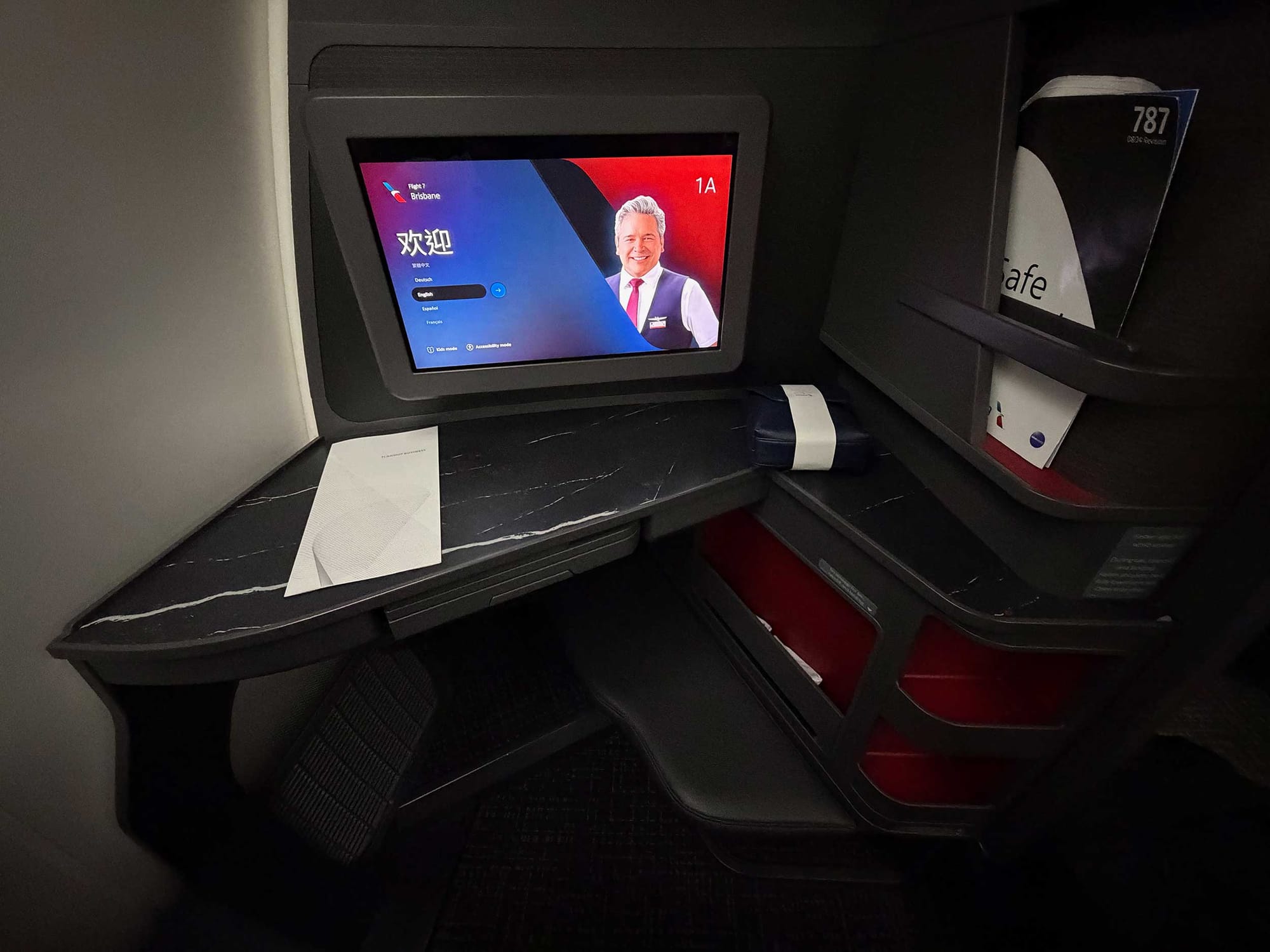 Wide angle view of entertainment screen, tabletop and storage cubbies with Boeing 787 safety card.
