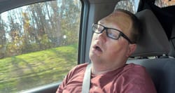 John asleep, with mouth open, in the front passenger seat of a mini van.