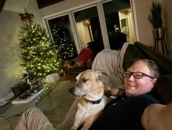 John lounging on the sofa with a dog, Christmas tree in the background.