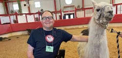 John seated in his wheelchair posing next to a llama in a barn.