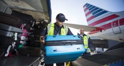 Baggage handler transferring suitcase from baggage cart into American Airlines airplane.