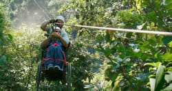 Person in wheelchair attached to zipline, going through forested area.