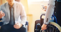 Two men in professional attire conversing in an office, one a wheelchair user.