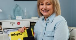 Blonde-haired woman wearing a light blue shirt, holding up a yellow tote with the word Adapts written on it.