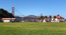 Green grassy field and historic buildings with Golden Gate Bridge in the background.