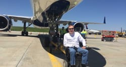 John seated in his wheelchair on the tarmac in front of a Boeing 757 airplane.