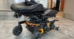 Permobil F3 power wheelchair with a bent wheel base, missing drive wheel and torn seat back and cushion.