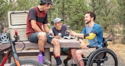 Wheelchair user sitting outdoors with two friends at a picnic table in a park.