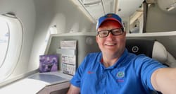 Selfie of John wearing blue polo shirt and hat seated in business class on a Fiji Airways airplane.