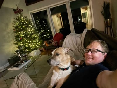 John lounging on the sofa with a dog, Christmas tree in the background.