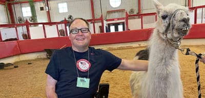 John seated in his wheelchair posing next to a llama in a barn.