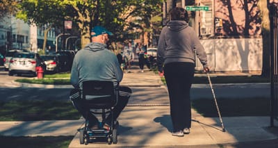 Woman with cane walking next to man using scooter on sidewalk.