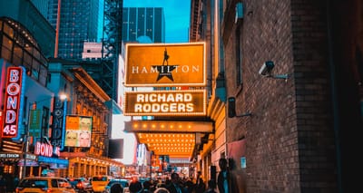 Hamilton play sign outside Broadway theatre on busy street.