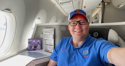 Selfie of John wearing blue polo shirt and hat seated in business class on a Fiji Airways airplane.