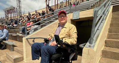 John seated in his wheelchair in a tunnel leading out onto the upper deck of seats at a stadium.