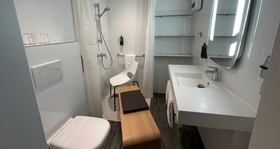 Hotel bathroom with a long wooden bench placed between narrow space and a plastic desk chair in the shower.