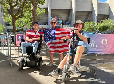 John seated in his wheelchair next to a man with a scooter and a woman holding a United States flag outside of a football sta