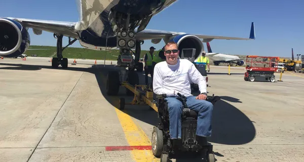 John seated in his wheelchair in front an airplane on the tarmac.