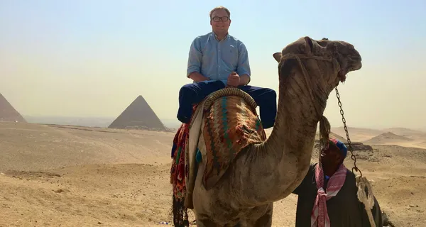 John seated on a camel in the Egyptian desert with pyramids in the background.