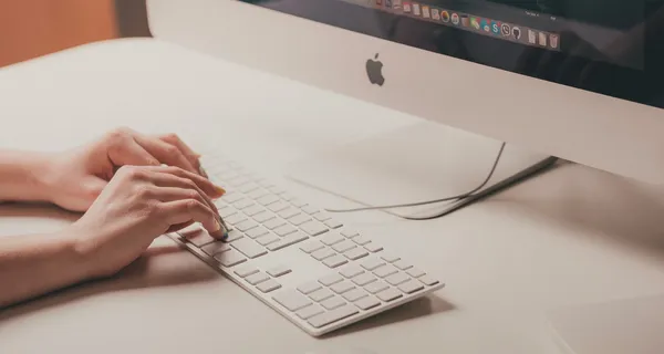Woman's hands resting on a keyboard in front of an Apple computer.