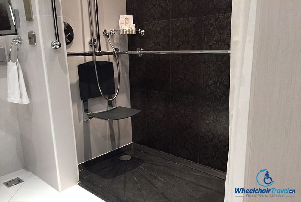 Photo Description: A roll-in shower with marble tile walls and floor, a shower seat attached to the wall, surrounded by grab bars.