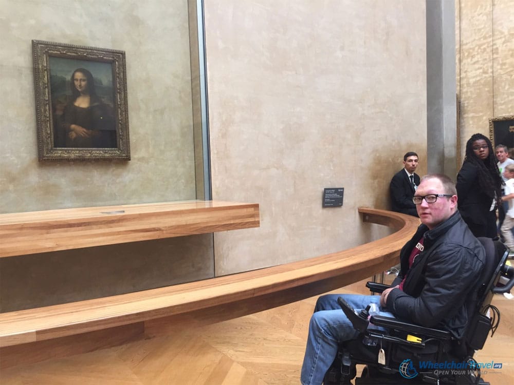 Photo Description: John Morris sitting in his power wheelchair, directly in front of the Mona Lisa at the Louvre Museum in Paris, France.