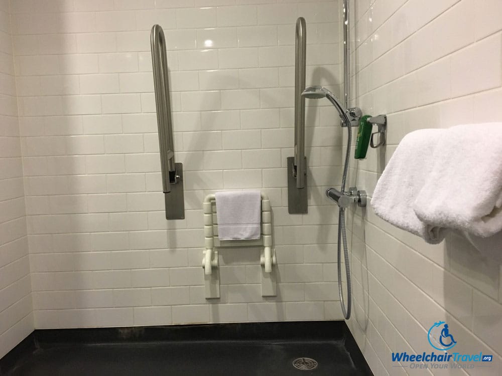 Photo Description: Roll-in shower with a shower seat attached to the wall, and two grab bars that can be folded up or down.