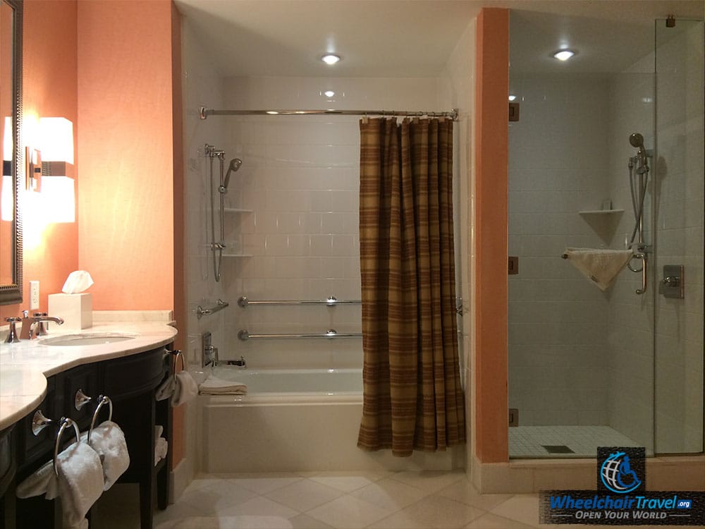 Photo Description: Wheelchair accessible hotel bathroom, with a sink/countertop, a tub with grab bars, and a walk in shower enclosed by glass.