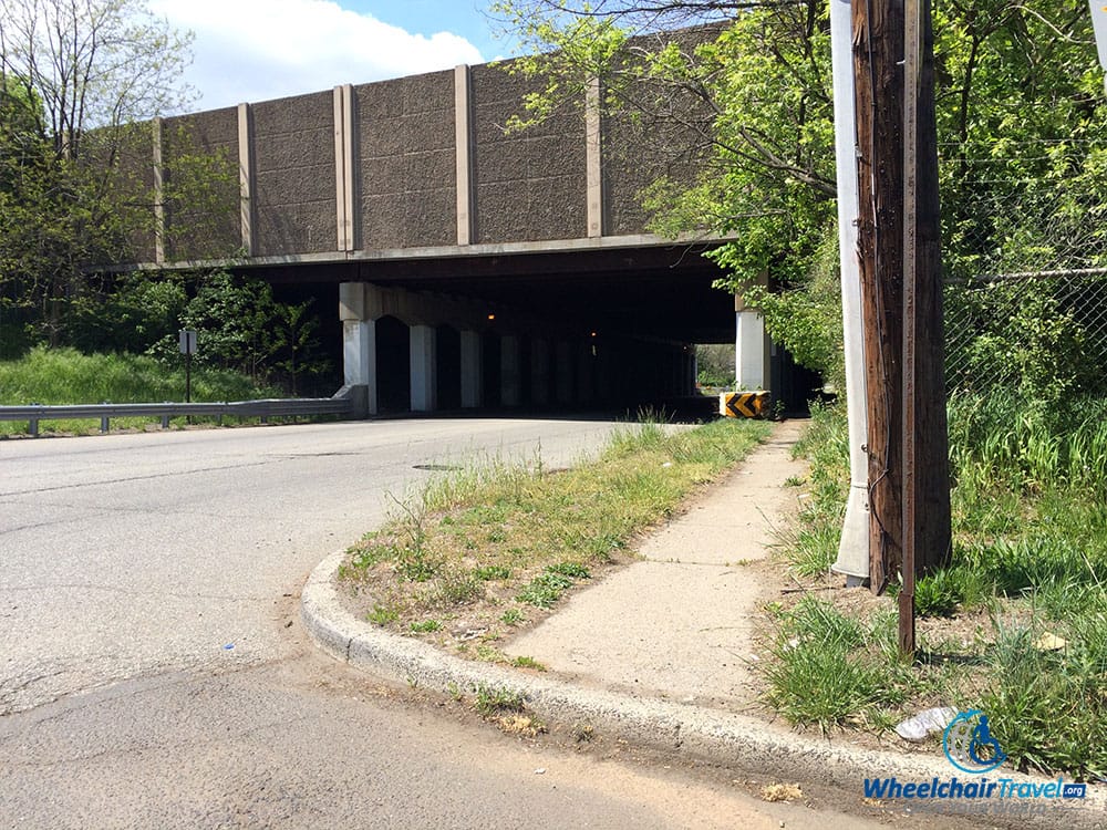 PHOTO DESCRIPTION: A sidewalk going under a dark highway overpass that has no accessible curb cut.