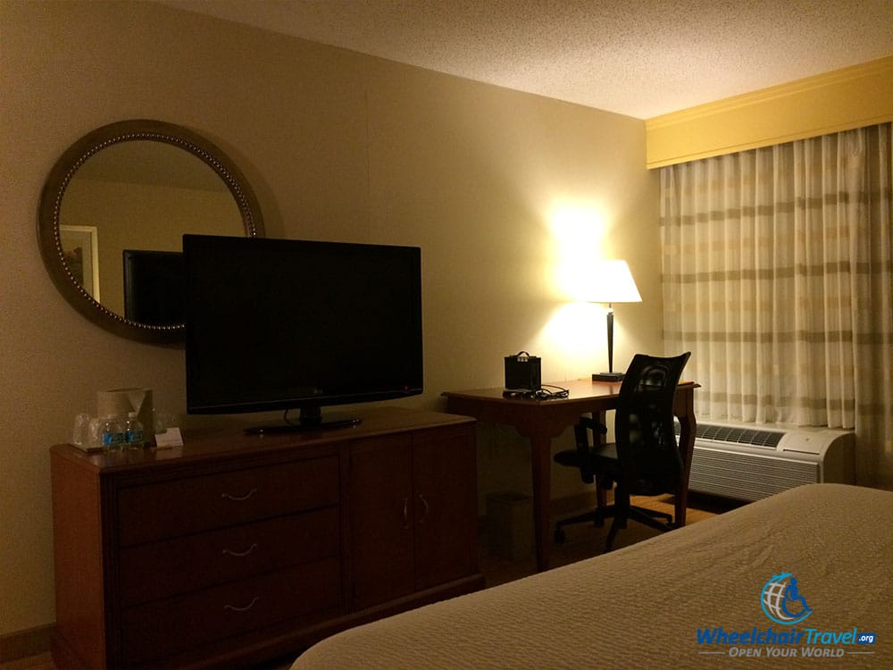 PHOTO DESCRIPTION: Looking across the bed to the opposite wall was a flat screen television atop a chest of drawers, and a desk with chair by the window.