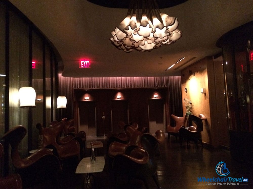 PHOTO DESCRIPTION: A dark lobby with 10 or more chairs & tables, with elevators across the room.