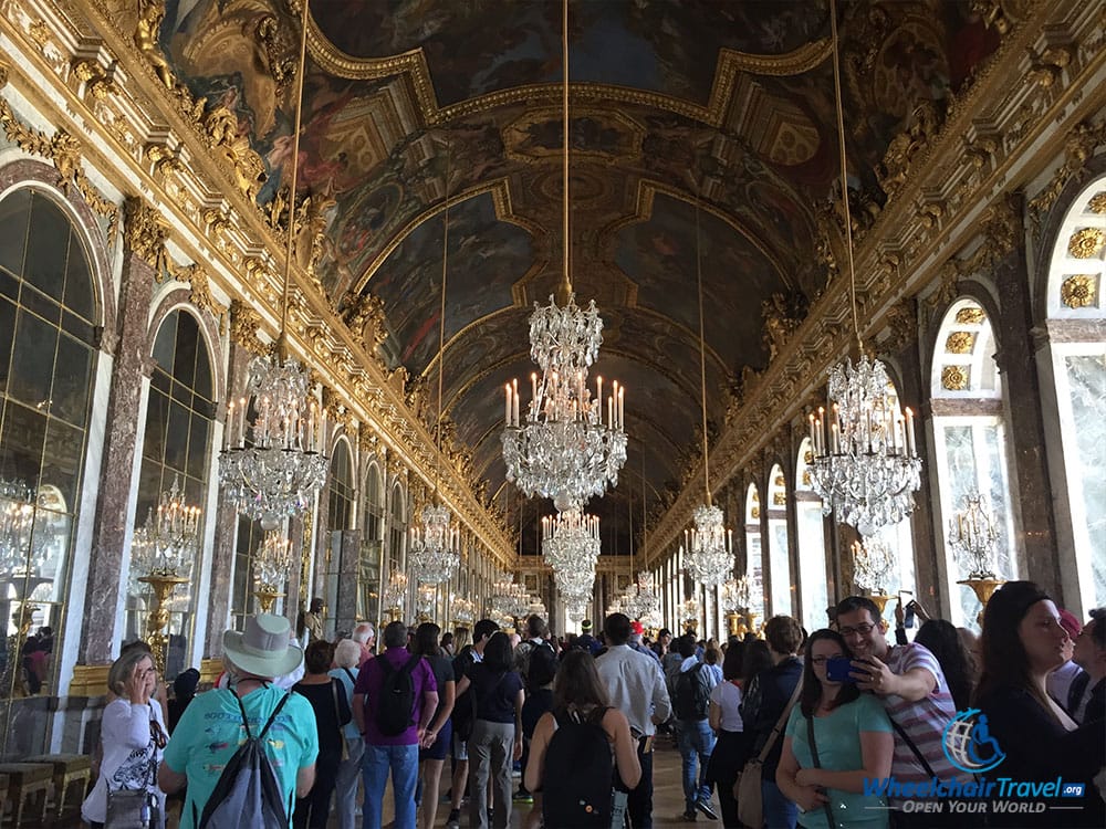 PHOTO DESCRIPTION: A long hall with mirrors on the left wall and windows on the right, glass and crystal chandeliers hang from the arched ceiling.