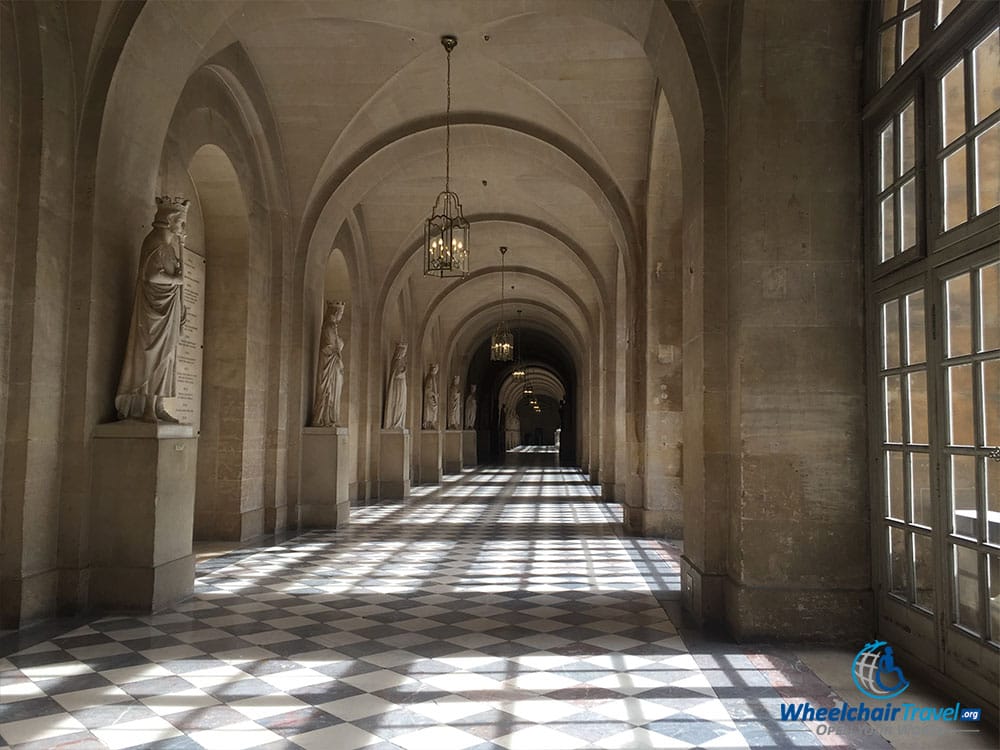 PHOTO DESCRIPTIONS: A long and empty hallway with windows on the right and pedestals topped with statues and busts on the left.