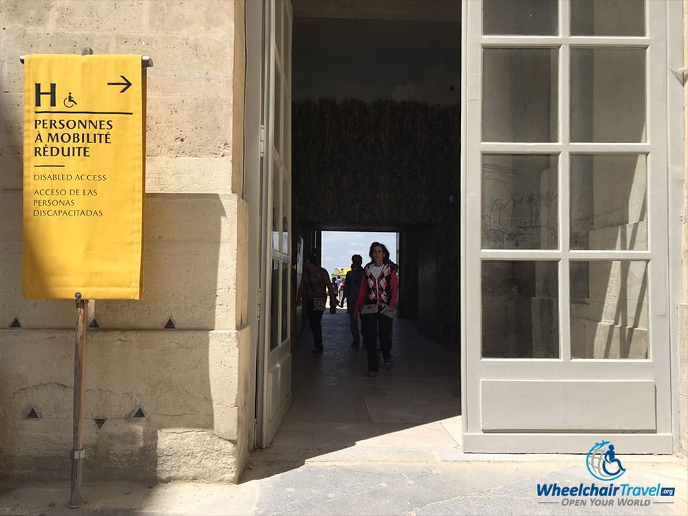 PHOTO DESCRIPTION: Open doorway into the palace, with a yellow banner outside the door that has a bolded letter H, a wheelchair icon and text that reads 'Disabled Access'.