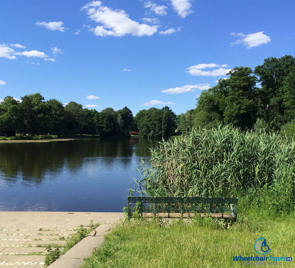 PHOTO DESCRIPTION: An empty bench surrounded by grass and weeds in front of a lake at Charlottenburg Palace.