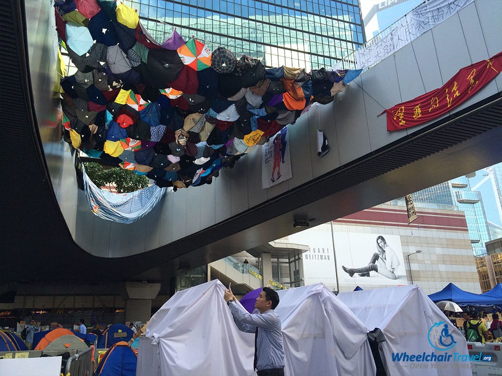 PHOTO DESCRIPTION: Man taking a photo on his mobile phone, will standing beneath a collection of umbrellas draped across a bridge.