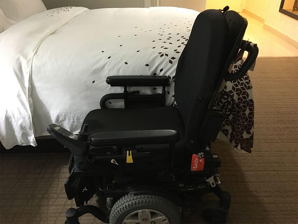 Power wheelchair parked next to hotel bed demonstrates significant height disparity.