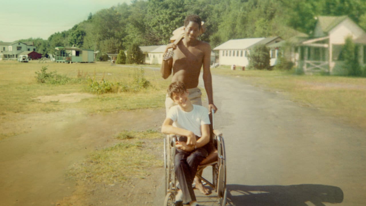 Historic photo of a man holding a guitar pushing another man with a wheelchair down a paved pathway of a summer camp.