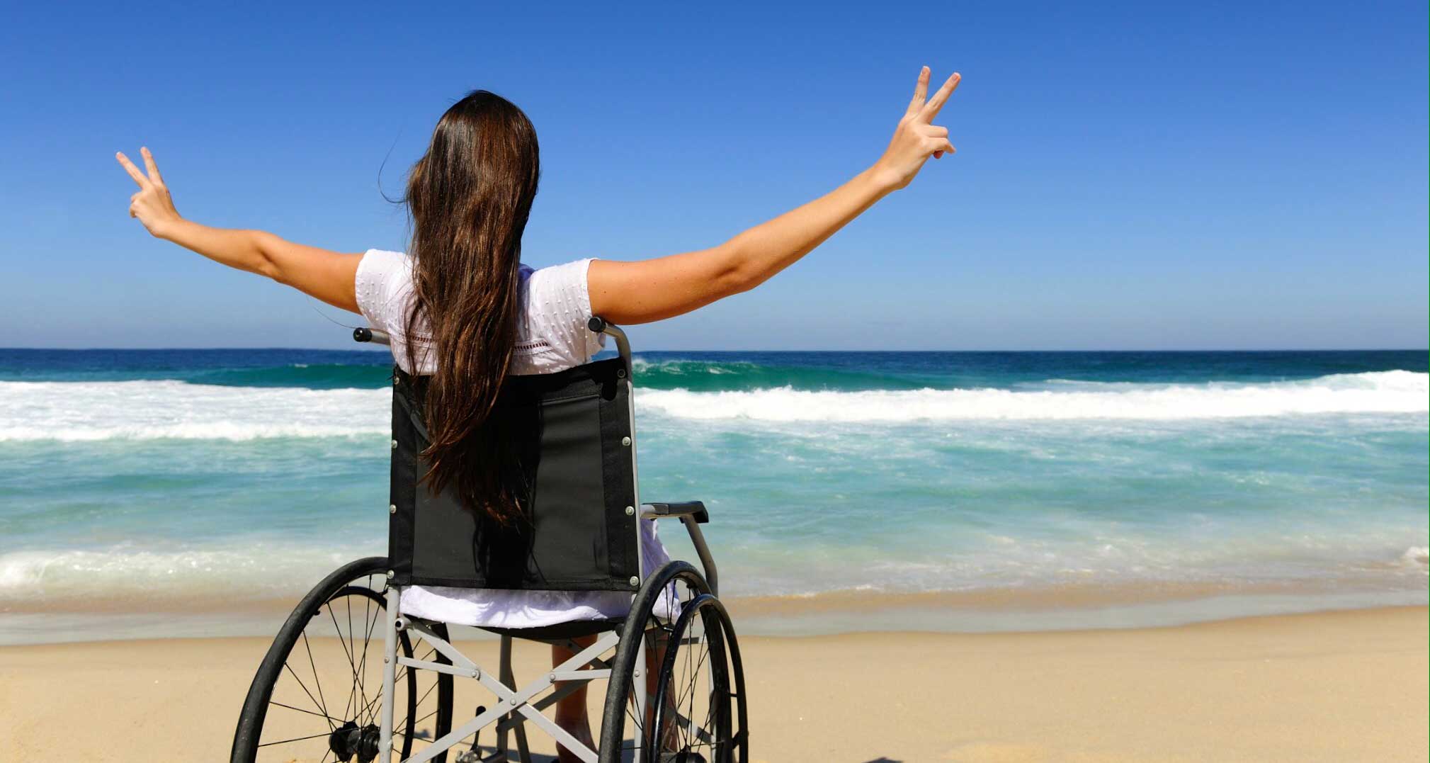 Stock photo of woman in wheelchair one beach with arms raised to heaven and hands making peace signs.