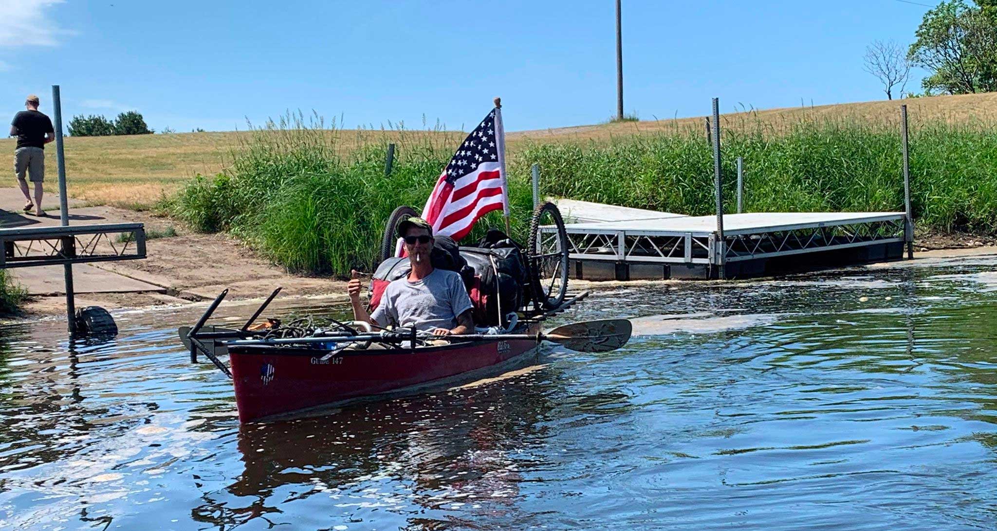 Man on canoe departing river launch point, with large American flag positioned at back of canoe.