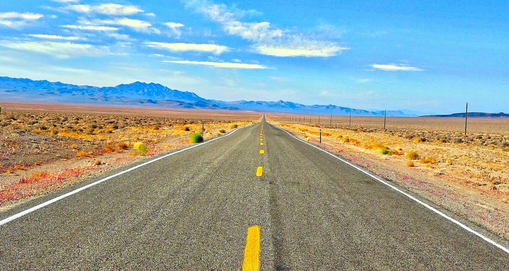 Two-lane road leading through desert-like area and toward mountains in the distance.